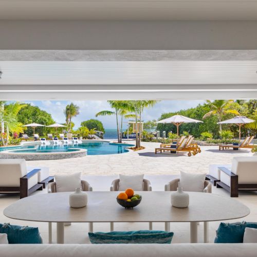 A tropical resort courtyard with a turquoise pool, sun loungers and umbrellas, palm trees, and ocean view beyond.