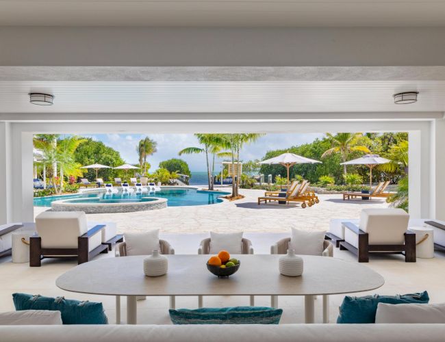 A tropical resort courtyard with a turquoise pool, sun loungers and umbrellas, palm trees, and ocean view beyond.