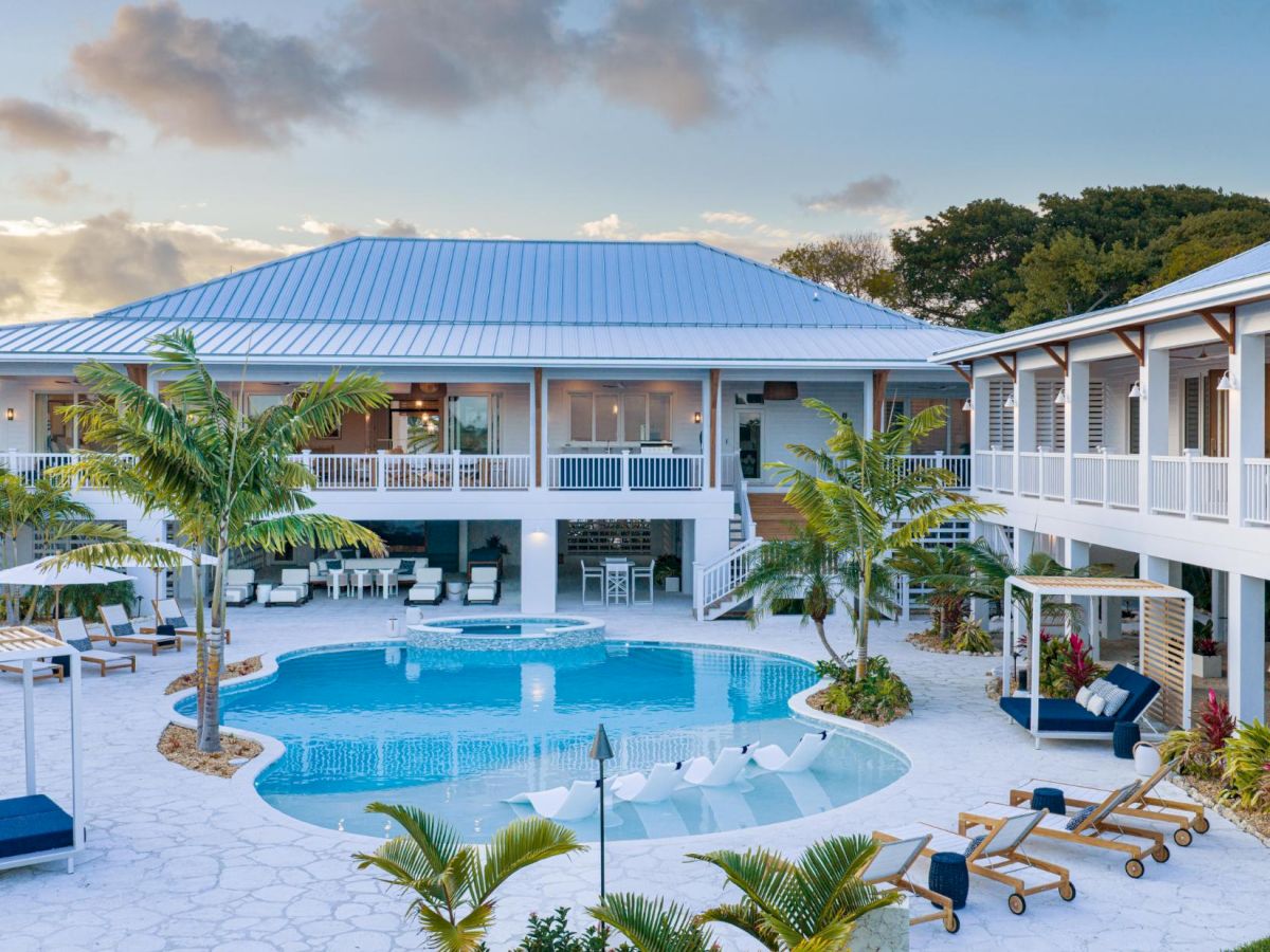 A tropical resort courtyard with a blue swimming pool, palm trees, lounge chairs, and white buildings with metal roofs.