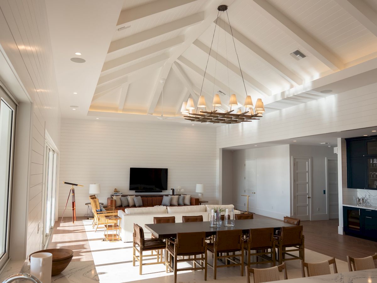 A bright, spacious open-plan living and dining area with a vaulted white ceiling, large windows, a long dining table, and a modern chandelier.