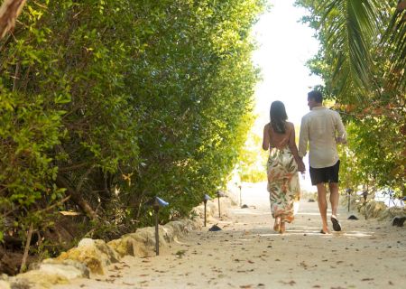 A couple walks hand-in-hand along a sandy path under lush green trees and bright sunlight.