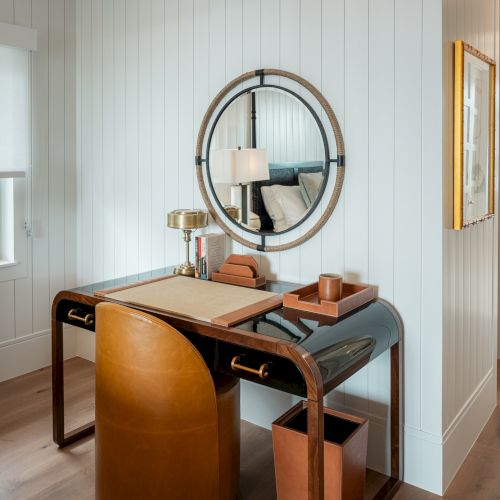 A vintage wooden desk with a swivel chair, a metal circular rack, a laptop, and a windowed room with soft natural light.