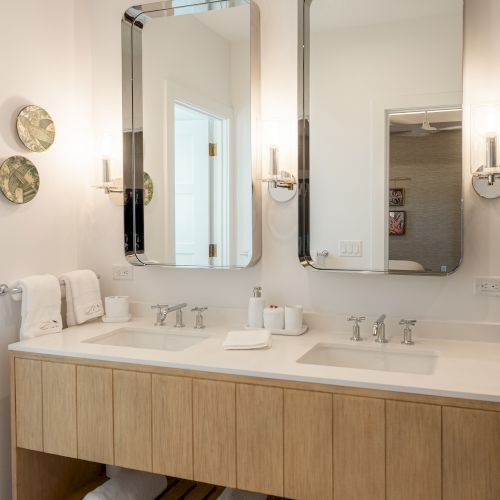 A modern bathroom vanity with two mirrors, wooden countertop, and an open shelf holding towels beneath; soft, neutral tones throughout.