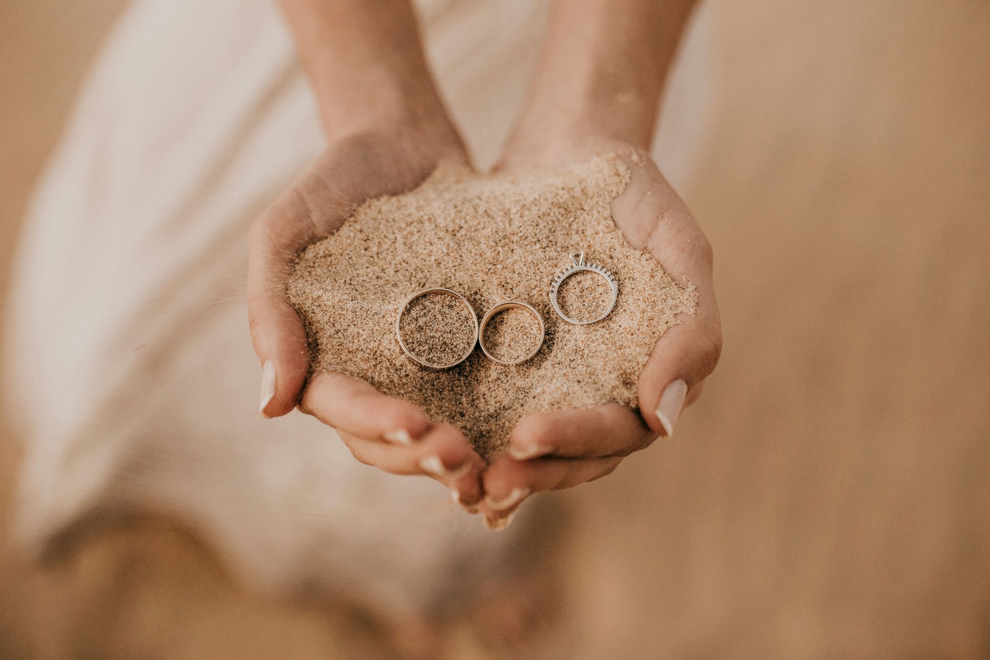 Hands are cupping a small pile of sand, with three rings partially buried in it.