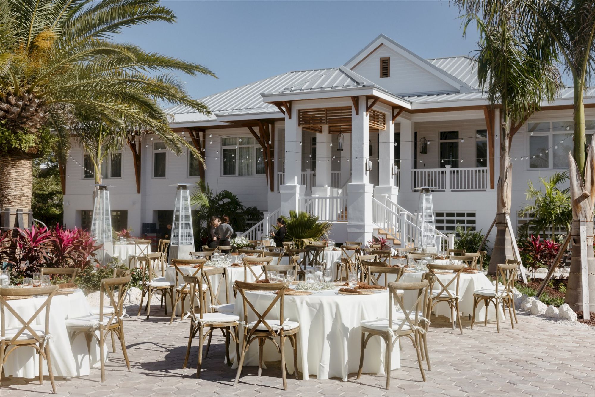 Outdoor wedding setup with round tables, white tablecloths, wooden chairs, and a charming house facade in the background.