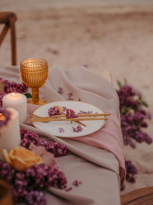 A beachside table setting with flowers, a candle, and a gold utensil set on a white plate, featuring a gold-rimmed glass.