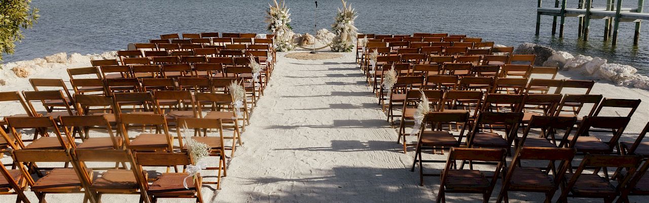 Beachside wedding setup with wooden chairs lining a sandy aisle, statues at the end, and a calm sea under a clear sky. Ends with a period.