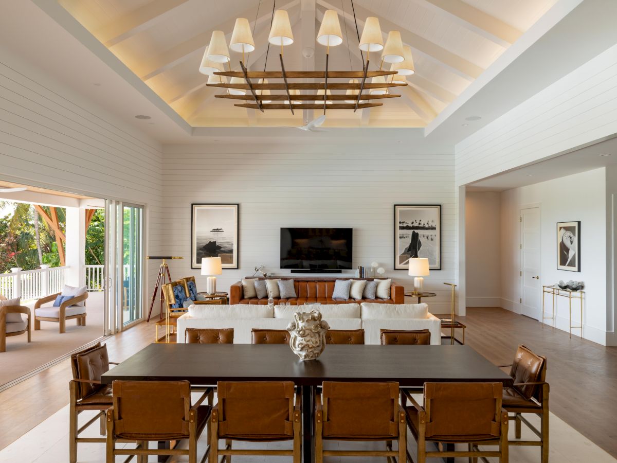 A spacious, modern living room with a high vaulted ceiling, white walls, panoramic glass doors, a large dining table, and a cozy seating area facing a TV.