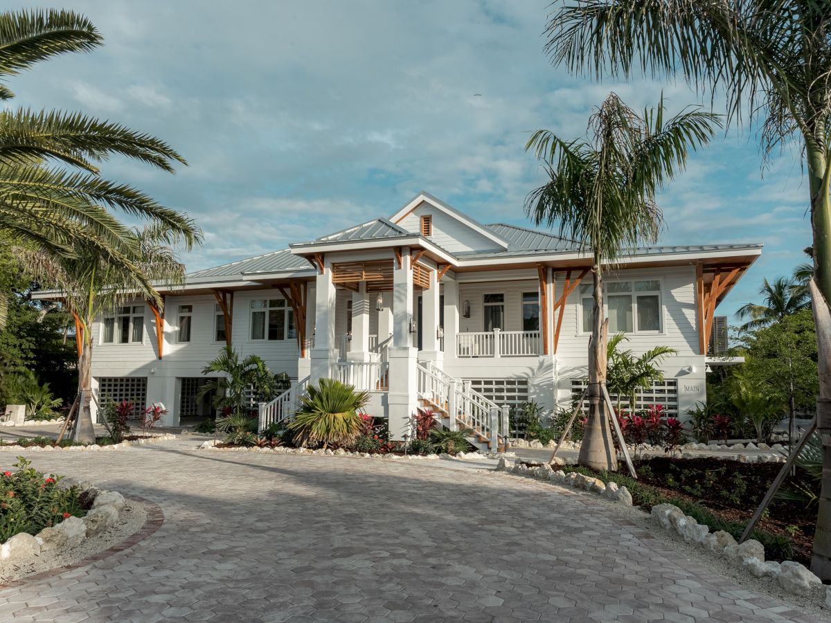 A large, white, single-family home with a curved driveway, palm trees, and tropical landscaping by the entrance, under a bright sky.