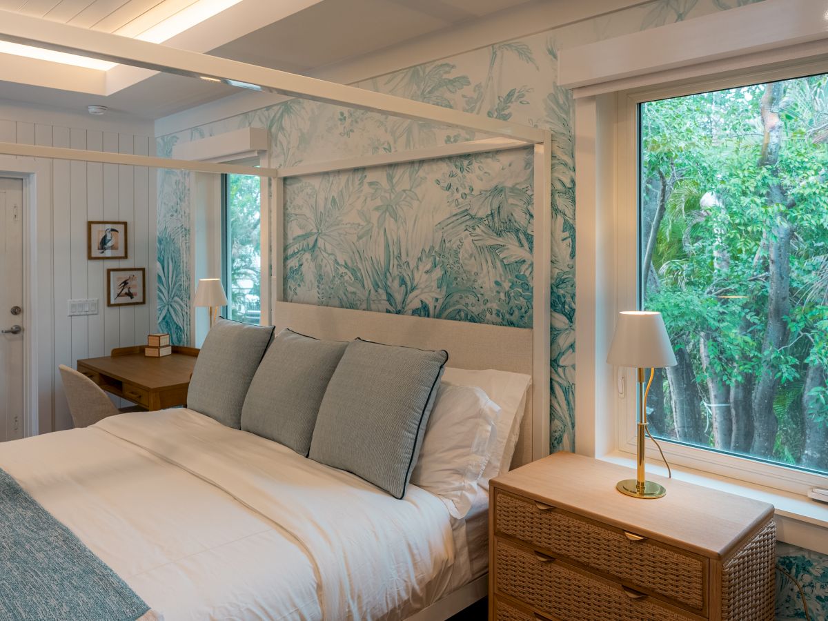 A cozy bedroom with a white-and-teal color scheme, a large bed with gray pillows, wicker nightstands, a desk near the door, and a big window showing green trees outside.