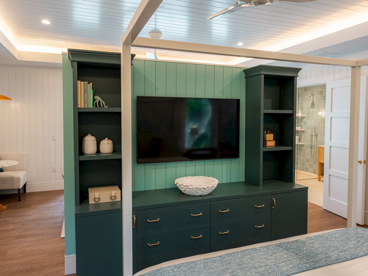 A teal entertainment wall with a mounted TV, cabinets and drawers, shelves with decor, and a light, cozy living area with a rug.