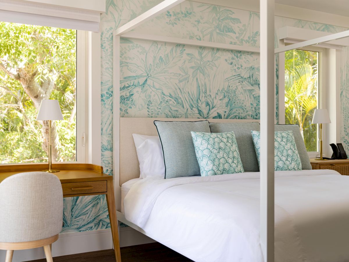 A bright, stylish hotel room with a four-poster bed, tropical wallpaper, white linens, teal throw pillows, and wooden desks by the windows, outdoorsy light.