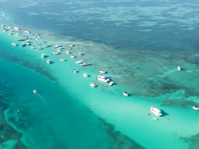 Aerial view of a tropical turquoise ocean with white sandy beaches and a small coastal village, bright sunny skies, and clear blue water.