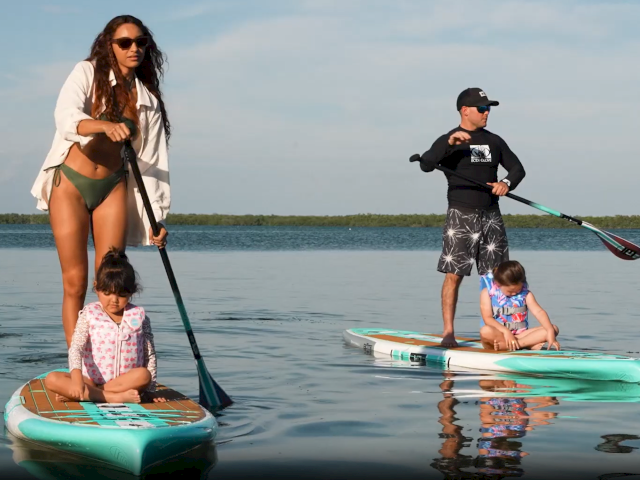 Three people on a sunny beach playing near the water, with two kids in swimsuits and an adult watching, waves rolling in.