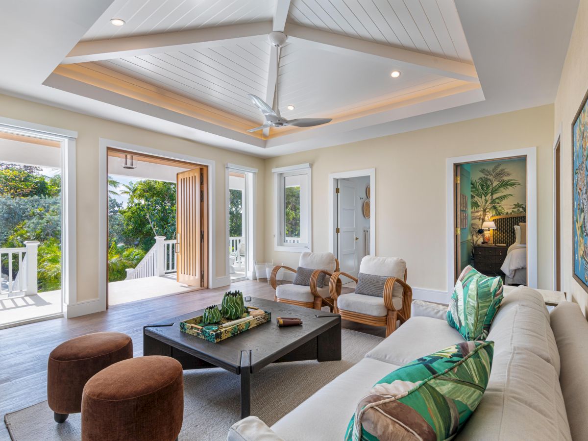 A bright, airy living room with beige walls, white sofas, green cushions, a dark coffee table, and a tray of decor; large windows and a patio door open to greenery, with a vaulted ceiling and ceiling fan.