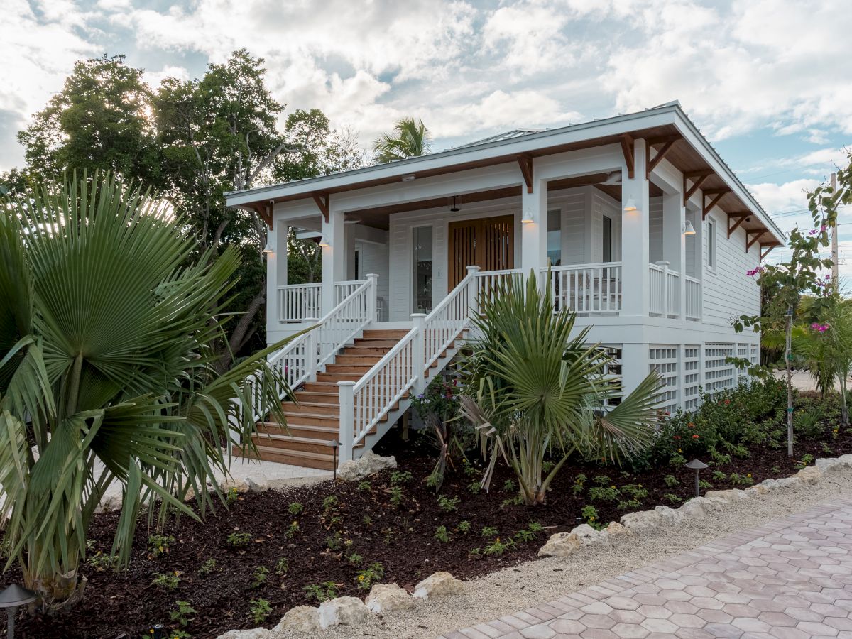 A white raised cottage with a surrounding porch, stairs to the front, and tropical plants along a stone path.