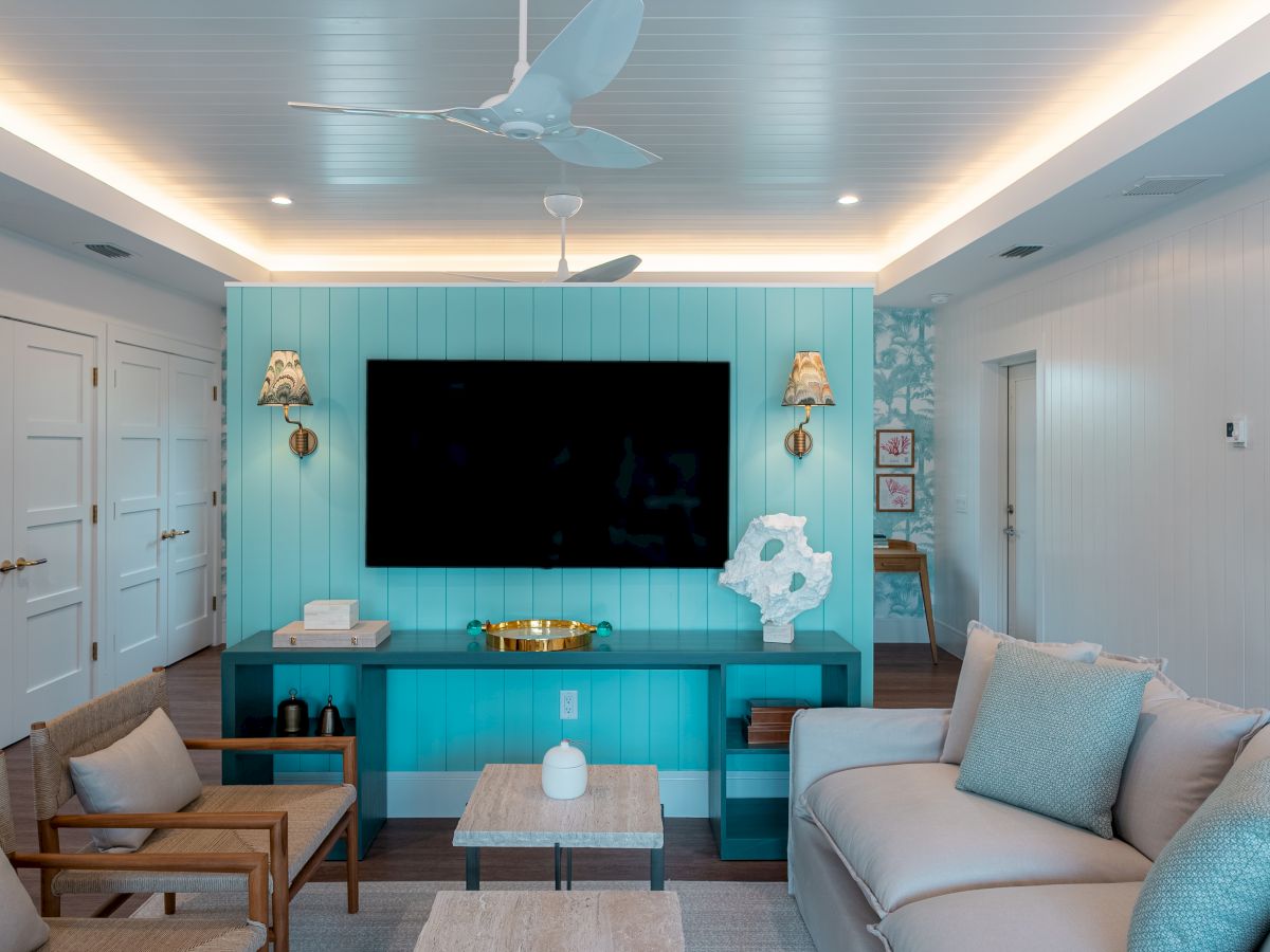 A bright living room with a turquoise feature wall, built-in entertainment unit, a large flat-screen TV, beige and teal seating, and coastal decor, ending with a cozy vibe.