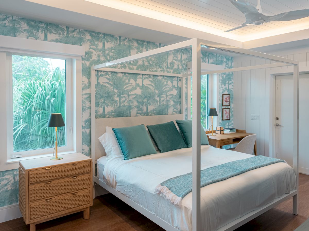 A stylish bedroom with a four-poster white bed, teal accents, tropical wallpaper, a wicker nightstand, and a desk area by the window, airy and bright.