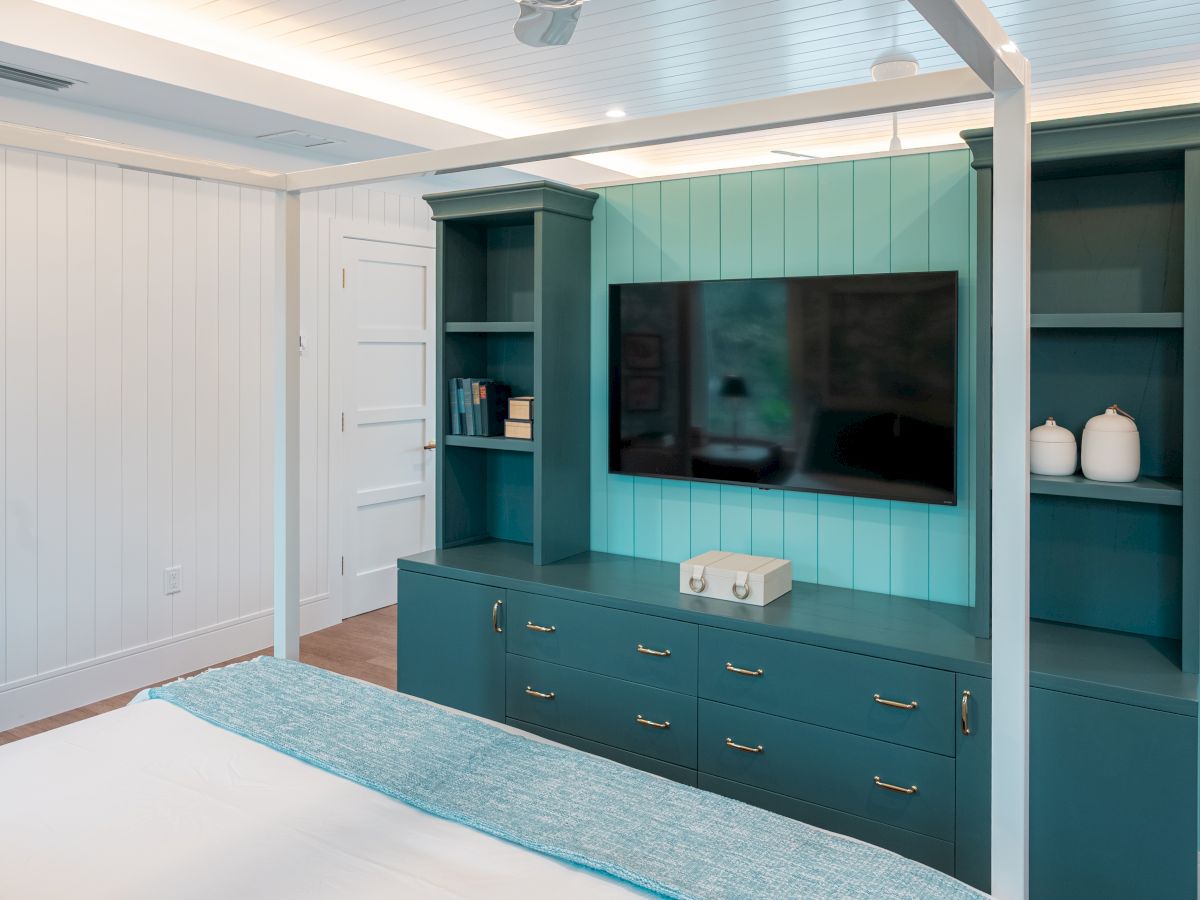 Tv wall unit with teal cabinetry, built-in shelves, a flat-screen TV, and a white storage box on top of the drawers in a bright, modern bedroom.