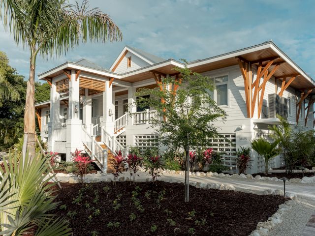 A modern single-story house with a white exterior, wooden support beams, a small front porch, palm trees, and landscaped garden under a blue sky.