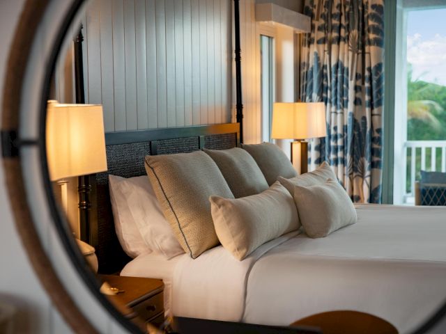 A cozy hotel bedroom with a made-up bed, throw pillows, lamps, and patterned curtains by a balcony.