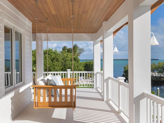 A white oceanfront balcony with a wooden porch swing, lantern lights, and sea views under a wooden ceiling.
