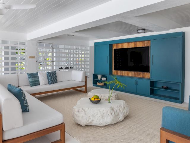 Bright living room with white sofa, blue media cabinet, flat-screen TV, and large white shutter windows.
