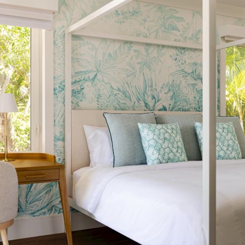 A bright bedroom with a neatly made bed, blue patterned pillows, bedside lamps, and large windows overlooking green trees.