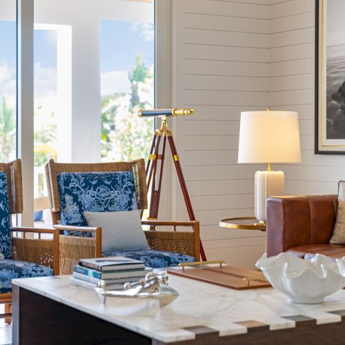 A bright living room with blue patterned chairs, a couch, a golden tripod lamp, and a marble coffee table near windows.