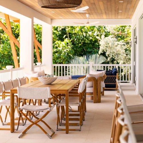 Sunlit outdoor patio with a wooden dining table, cushioned chairs, and a view of lush tropical greenery.
