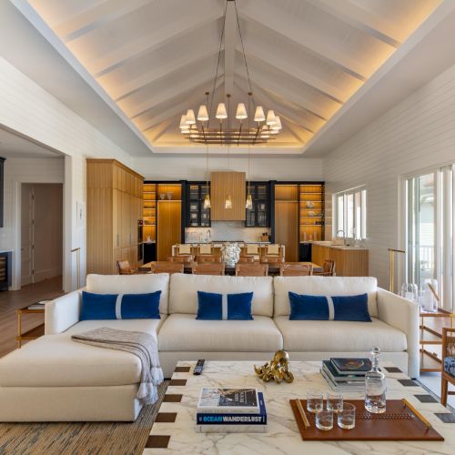 Bright modern great room with white walls, wood cabinetry, a chandelier, a large sofa, and a dining table opening to patio doors.
