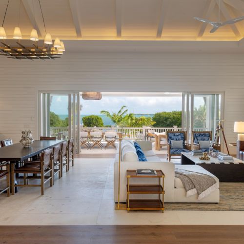 Bright modern living and dining area with sofas, coffee tables, a chandelier, and open doors to a sunny ocean view.
