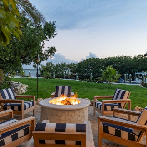 A cozy outdoor fire pit with lounge chairs sits in a tropical garden overlooking the ocean under a partly cloudy sky.