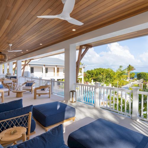 Sunlit tropical veranda with cushioned seating, wooden ceiling and ceiling fan, overlooking palm trees and the ocean.