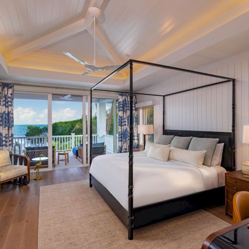 A bright ocean-view bedroom with a four-poster bed, ceiling fan, patterned curtains, and soft lamps by large windows.