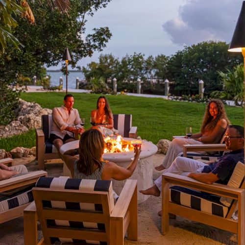 Friends relax on a seaside lawn around a lit fire pit, toasting with drinks as warm lights glow among the trees.