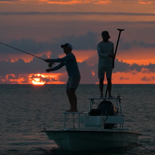 Two anglers stand on a small boat fishing in the ocean at sunset under colorful clouds.