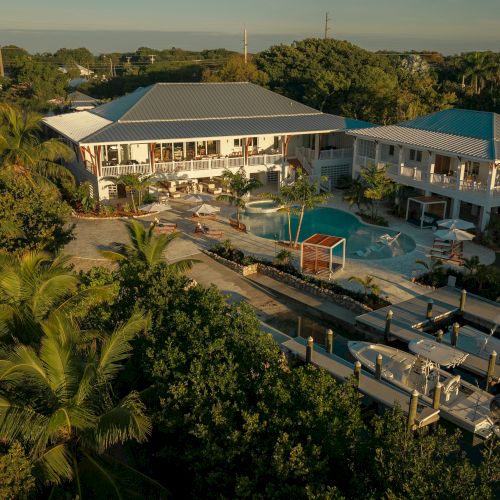 Aerial view of a tropical resort with a turquoise pool, sun loungers, palm trees, and white buildings under a blue sky.