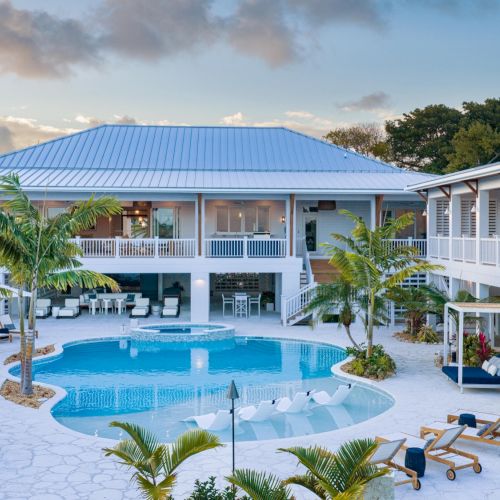 A tropical resort courtyard with a blue swimming pool, white lounge chairs, palm trees, and modern villas with blue metal roofs.