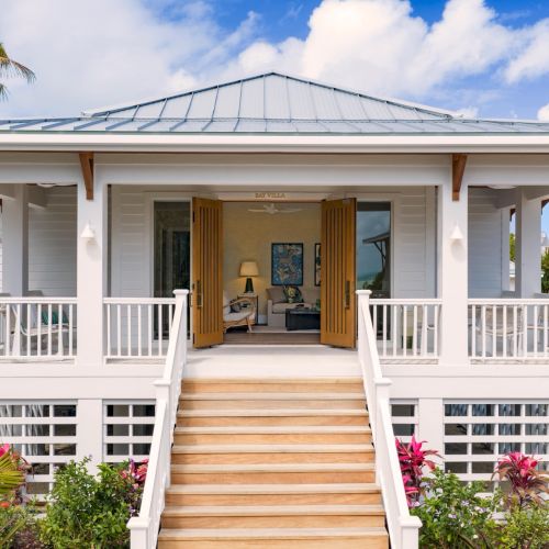 A modern white tropical villa with a covered porch, wooden steps, open doors, palm trees, and blue sky with clouds.