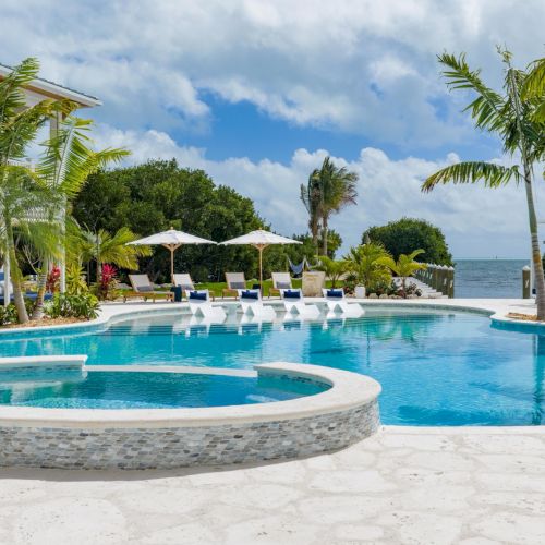 A tropical beachfront resort with a large blue pool, loungers under umbrellas, palm trees, and the ocean beyond.