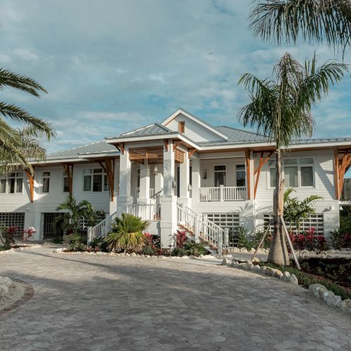 A bright white modern house with a covered porch, surrounded by tropical palm trees and landscaped gardens under a blue sky.