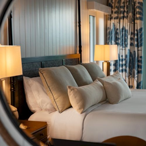 A hotel-style bedroom with a made bed, pillows, bedside lamps, curtains, and a view of greenery outside the window.