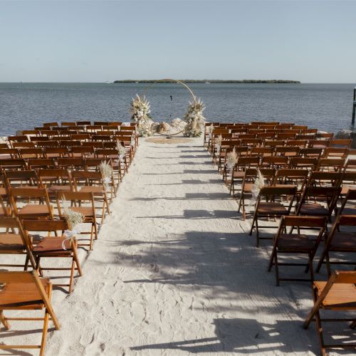 A beach wedding setup with rows of wooden chairs, a floral aisle arch, and the ocean in the background.