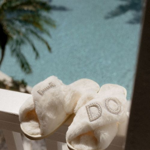 White plush heart slippers with pearl letters sit by a window, with a small ring-like charm on the left toe.