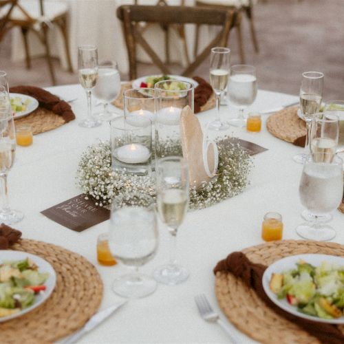 An outdoor brunch/celebration table set with plates of salad and bread, many water glasses, candles, and tea lights, ready for guests.