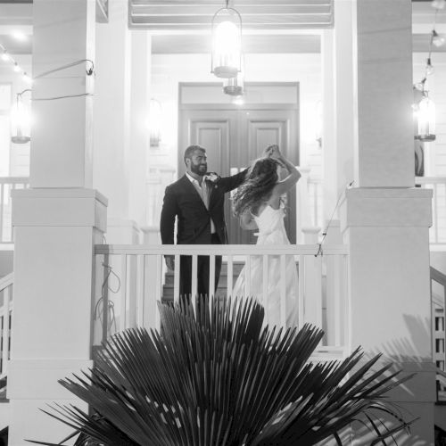 A bearded man in a suit helps a woman with her dress inside a bright hall with chandeliers and railings.