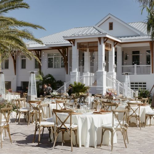 An outdoor reception setup with a decorated table, chairs, string lights, and a modern white house surrounded by palm trees under sunny skies.